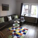 3Sitzer-Couch mit Recamiere, bunter Teppich, zwei Tablett-Tischchen, Laminatboden, graue Gardinen